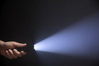 Black flashlight with wide beam in male's hand isolated from left side of the frame on black background