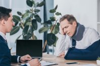 upset worker in neck brace with broken arm sitting at table opposite businessman in blue jacket in office, compensation concept