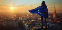 A woman wearing the flag of the European Union e is standing on top overlooking a city during the golden hour