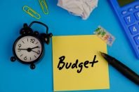 handwritten of BUDGET on yellow sticky note with black alarm clock and calculator on blue background. budget means to make a plan for the amount of money to spend on something. business concept.