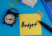 handwritten of BUDGET on yellow sticky note with black alarm clock and calculator on blue background. budget means to make a plan for the amount of money to spend on something. business concept.