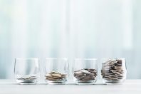 Glass jars filled with coins are arranged in ascending order on a wooden table against a bright white background. The image symbolizes the concept of financial growth and saving money over time.