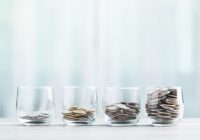 Glass jars filled with coins are arranged in ascending order on a wooden table against a bright white background. The image symbolizes the concept of financial growth and saving money over time.