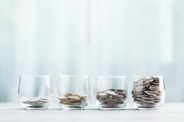 Glass jars filled with coins are arranged in ascending order on a wooden table against a bright white background. The image symbolizes the concept of financial growth and saving money over time.