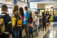 Students standing in line at a school cafeteria, waiting to get their lunch. They carry backpacks and engage in casual conversation.