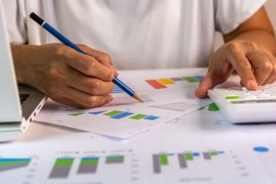Hand writes down something while using calculator, sitting at the laptop at home for study, audit, remote work, accountant, calculator, count, business, work from home concept.