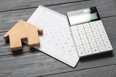 Wooden house figure, calendar and calculator on grey wooden background. Debt concept