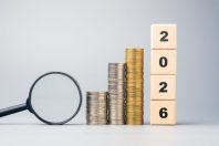 Stacked coins, magnifying glass, and wooden blocks showing 2026, symbolizing future financial goals, investment planning, and economic outlook for the coming year