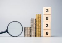 Stacked coins, magnifying glass, and wooden blocks showing 2026, symbolizing future financial goals, investment planning, and economic outlook for the coming year