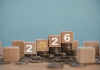 Wooden cube with the year 2026 on top of a stack of coins, symbolizing investment strategies, financial planning, income goals, growth, action and success for the new year.