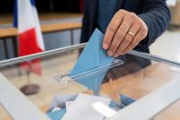 Close-up of a voter's hand casting a ballot into a transparent box during an election in France. Blue envelope being inserted with a blurred French flag in the background at a polling station.
