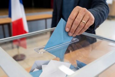 Close-up of a voter's hand casting a ballot into a transparent box during an election in France. Blue envelope being inserted with a blurred French flag in the background at a polling station.